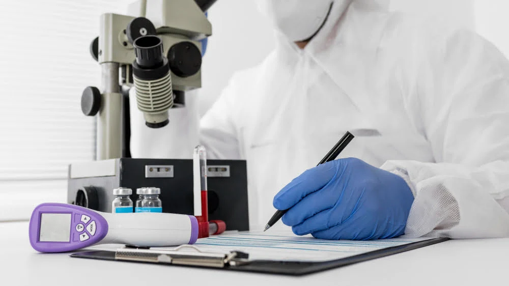 lab technician in protective gear writes on a clipboard with a microscope and vials in the background inside a lab.