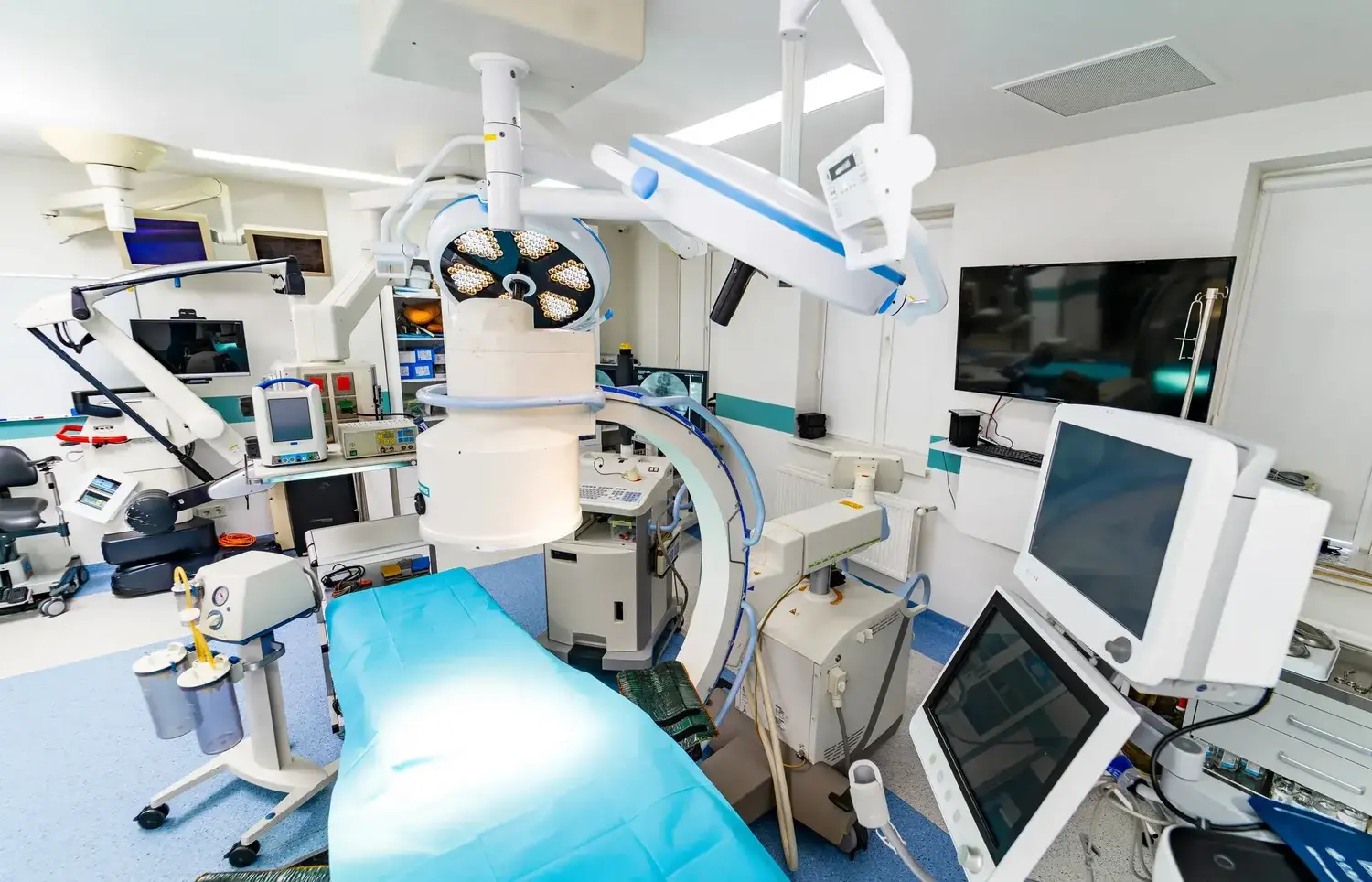 operating room with a blue-draped surgical table, overhead lights, and multiple monitors and anesthesia equipment surrounding the space.